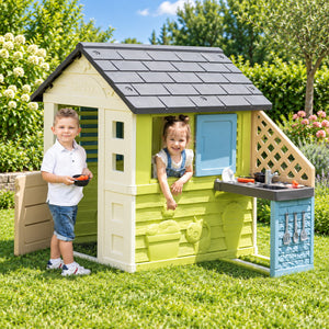 Deux enfants jouant dans et autour de la maisonnette Smoby Maison Pretty dans un jardin
