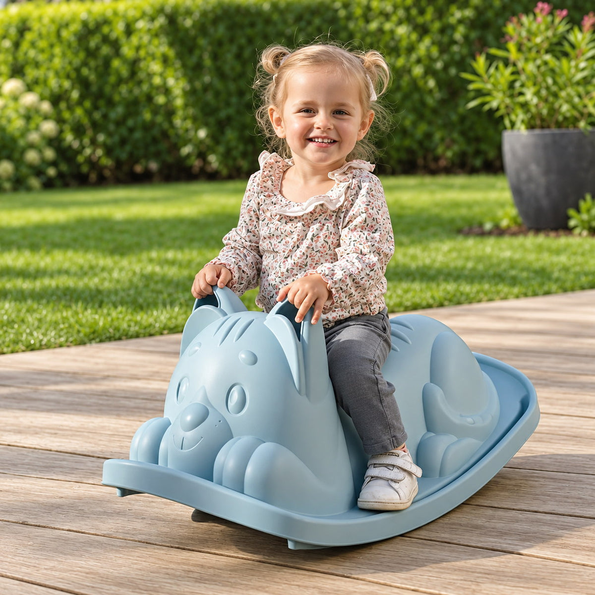 Petite fille assise sur la bascule chat Smoby Life bleue, installée sur une terrasse en bois dans un jardin